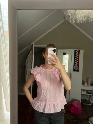 Rosa volangblus med broderi - Söt ljusrosa blus med volangdetaljer och broderade hålmönster. Blusen har rund halsringning, korta volangärmar och en lös passform med volang nedtill. Perfekt för en romantisk och feminin stil.