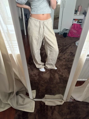 Beiga vida linnebyxor med dragsko - Säljer ett par beiga linnebyxor med avslappnad passform och dragsko i midjan. Byxorna har raka ben och elastisk midja för extra komfort. Perfekta för en chill och stilren look. Materialet är luftigt linne som känns svalt och skönt.