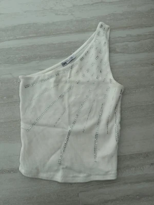 Vit oneshoulder topp från Zara - Säljer en vit oneshoulder topp från Zara med silverfärgade paljettdetaljer i ett coolt mönster. Toppen är ribbad och har en unik axelbandslös design som ger en trendig vibe. Perfekt för dig som vill sticka ut lite extra.