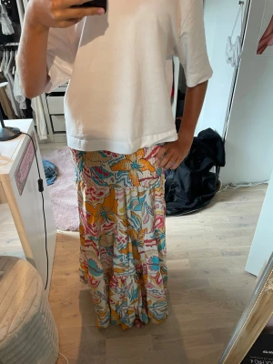 Färgglad långkjol med blommigt mönster - Säljer en färgglad långkjol med blommigt mönster i gult, rosa, blått och vitt. Kjolen har en flowy passform och resår i midjan för extra komfort. Perfekt för dig som vill sticka ut med en unik och somrig stil.
