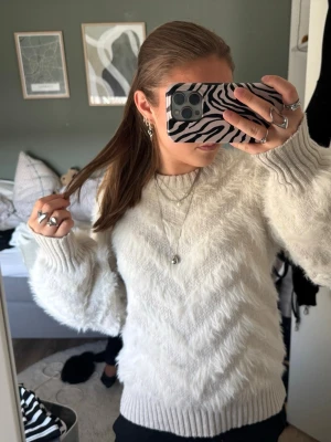 Vit fluffig stickad tröja - Mysig vit stickad tröja med fluffig textur och diskret chevronmönster. Tröjan har rund halsringning, långa ärmar och breda ribbade muddar vid ärmslut och nederkant. Perfekt för kalla dagar och ger en trendig look.