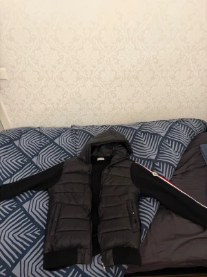 Svart monclercardigan - Snygg svart dunväst med huva och dragkedja framtill. Västen har två fickor med dragkedja och quiltad design. Perfekt för lager-på-lager och funkar till många olika stilar. Klassisk look med sportig touch.