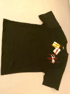 Corteiz  - Svart t-shirt med vit och röd grafisk print på både fram- och baksidan. T-shirten har korta ärmar och en normal passform. Detaljer med vita linjer på ryggen och tryck framtill ger en cool streetkänsla.
