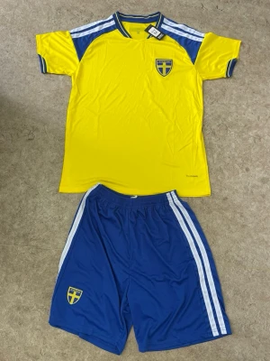 Svenskt fotbollsställ gul/blå - Säljer ett svenskt fotbollsställ med gul t-shirt och blå shorts. Tröjan har blå detaljer på axlar och ärmar samt Sveriges landslagsemblem på bröstet. Shortsen är blå med vita ränder och har även landslagsemblem. Tillverkat i lätt och snabbtorkande material.