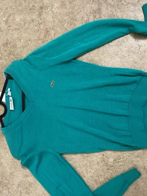 Turkos stickad tröja från Lacoste - Snygg turkos stickad tröja från Lacoste med klassisk rund halsringning och det ikoniska krokodilmärket broderat på bröstet. Tröjan har långa ärmar och ribbade muddar vid ärmslut och nederkant. Perfekt för dig som gillar stilrena plagg.