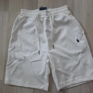 Vita shorts med snörning - Snygga vita shorts med elastisk midja och snörning. Klassisk design med två sidofickor och en liten broderad logga på vänster ben. Perfekta för varma dagar och enkel att matcha med olika stilar.
