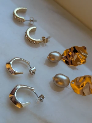 Tre par örhängen i silver och guld - Säljer tre par örhängen: ett par silvriga hoops med små stenar, ett par chunky silvriga hoops och ett par guldiga örhängen med oregelbunden form och hängande pärlor. Perfekta för att mixa och matcha till olika looks. De långa till höger vet jag ej vart jag köpte de ifrån, de andra två paren är från KappAhl 