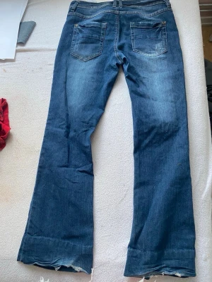 Blå bootcut jeans från KappAhl - Säljer ett par blå bootcut jeans från KappAhl med slitningar framtill och råa kanter vid bensluten. Jeansen har två fickor fram och två bak, samt en klassisk femficksdesign. Materialet är denim i bomull med lite stretch för skön passform.