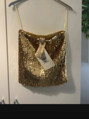 Guldig paljett tubtopp med smala band - Säljer en glittrig guldig tubtopp med smala axelband och paljetter över hela toppen. Perfekt för dig som vill sticka ut och glänsa. Materialet är en mix av polyvinyl, akryl och spandex vilket gör den stretchig och bekväm.