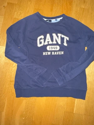 Mörkblå sweatshirt från GANT - Mörkblå sweatshirt från GANT med tryck 'GANT 1949 NEW HAVEN' på bröstet. Klassisk rund halsringning och långa ärmar. Tillverkad i mjuk bomullsmix för en avslappnad och sportig look.