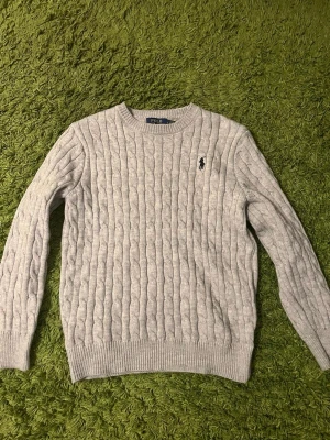 Ljusgrå kabelstickad tröja Polo Ralph Lauren - Kabelstickad tröja i ljusgrått från Polo Ralph Lauren med rund halsringning och broderad logga på bröstet. Tröjan har ribbade muddar vid ärmslut och nederkant, perfekt för lager-på-lager och en clean look.
