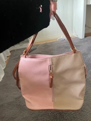 Tvåfärgad handväska i rosa och beige - Snygg handväska i rosa och beige konstläder med bruna axelremmar och guldfärgade detaljer. Väskan har dragkedja framtill och baktill samt justerbara remmar. Perfekt för dig som gillar en stilren men lekfull look.