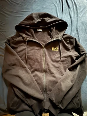 Svart EA7 hoodie med dragkedja - Svart hoodie från EA7 Emporio Armani med dragkedja framtill, justerbar huva och fickor på sidorna. Liten gul EA7-logga på bröstet och gul detalj på ryggen. Tillverkad i mjukt bomullsmaterial, perfekt för en avslappnad streetwear-look. Ser solbränd ut på bilderna men den är helt ny