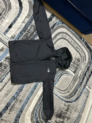 Svart regnjacka från The North Face - Snygg svart regnjacka från The North Face med huva och dragkedja framtill. Jackan har en minimalistisk design, logga på bröstet och är tillverkad i ett vattenavvisande material. Perfekt för regniga dagar när du vill hålla dig torr.