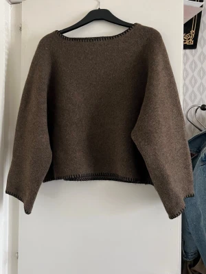 Brun stickad tröja med kontrastsömmar – oversize - Snygg stickad tröja i melerad brun nyans med dekorativa svarta kontrastsömmar längs halsringning, ärmslut och nederkant. Mjuk och bekväm passform med lätt oversize-känsla. Perfekt till höst och vinter.