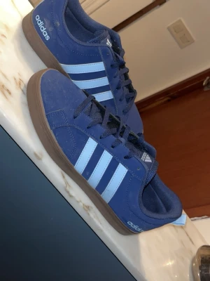 Blå Adidas sneakers med ljusblå ränder - Snygga blå sneakers från Adidas med klassiska ljusblå ränder på sidorna och brun sula. Skorna har lågt skaft, snörning och är tillverkade i mockaliknande material. Perfekta för en avslappnad streetstyle-look.