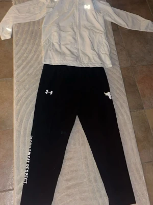 Under Armour träningsset svart/vit - Säljer ett träningsset från Under Armour med en vit zip-jacka och svarta byxor. Byxorna har texten 'Blood Sweat Respect' längs benet och logga på låret. Jackan har dragkedja och logga på bröstet. Perfekt för gym eller löpning.