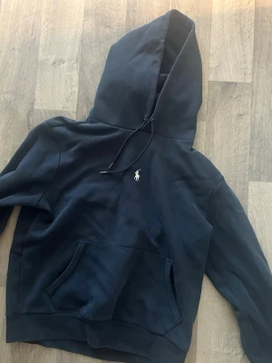Mörkblå hoodie från Polo Ralph Lauren - Snygg mörkblå hoodie från Polo Ralph Lauren med klassisk vit logga på bröstet. Hoodien har stor huva med dragsko och en rymlig magficka. Tillverkad i mjukt bomullsmaterial som känns skönt mot huden. Perfekt för en avslappnad och stilren look.