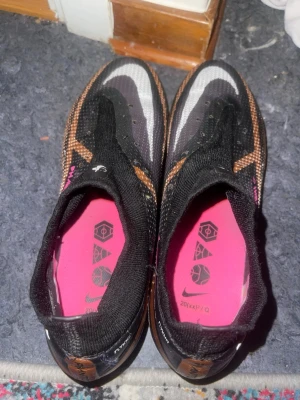 Nike Phantom GT2 fotbollsskor svart/orange - Säljer ett par Nike Phantom Elite GT2 fotbollsskor i svart med orange och vita detaljer. Skorna har strumpliknande skaft, rosa innersula och tydliga loggor. Ovandelen är i syntetmaterial med textur för bättre bollkontroll. Perfekta för dig som vill sticka ut på planen.