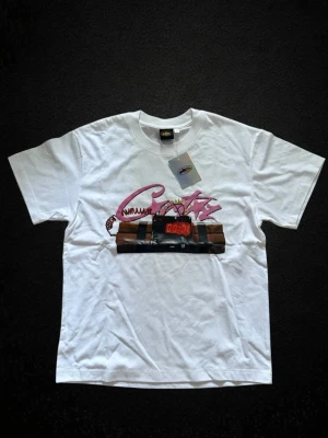 Vit t-shirt med tryck från corteiz  - Säljer en vit t-shirt från Adieuite i storlek M. T-shirten har ett färgstarkt tryck framtill med rosa text och en digital klocka. Klassisk passform och rund halsringning. Perfekt för dig som vill sticka ut med en unik design.