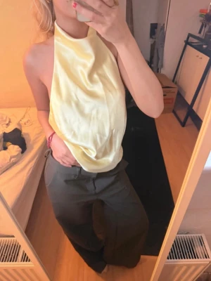 Gul halterneck topp i satin - Säljer en ljusgul halterneck topp i glansigt satinmaterial. Toppen är ärmlös och har öppen rygg, vilket ger en snygg och trendig look. Perfekt för dig som vill sticka ut med en unik och fräsch stil.