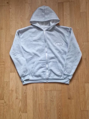 Ljusgrå zip-hoodie från Corteiz - Ljusgrå hoodie med dragkedja från Corteiz. Stor logga i vitt på ryggen och mindre tryck på bröstet. Utrustad med huva och känguruficka framtill. Mjuk bomullskänsla och ribbade muddar vid ärmslut och nederkant. Storlek S men passar även på M