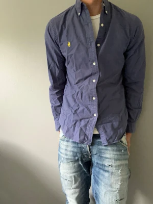 Marinblå skjorta från Ralph Lauren - Klassisk marinblå skjorta från Ralph Lauren med knappkrage och vit knappstängning. Skjortan har lång ärm och den ikoniska gula logotypen broderad på bröstet. Tillverkad i bomull och har en normal passform som funkar till många olika stilar.