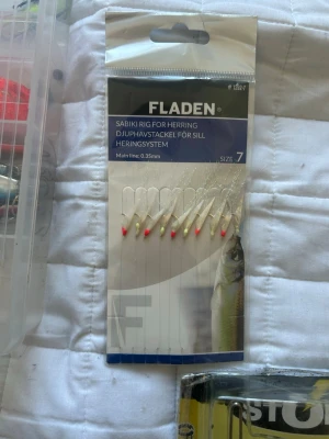 Fladen silltackel storlek 7 - Djuphavstackel för sill från Fladen, modell Sabiki Rig for Herring i storlek 7. Huvudlina 0,35 mm. Flera krokar med vita och röda detaljer, perfekt för fiske efter sill. Förpackningen är genomskinlig med blå och vita detaljer.
