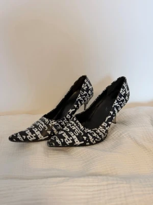 Svarta pumps med vit textprint - Säljer ett par svarta pumps med spetsig tå och vit textprint över hela skon. Klackarna är smala och i metall, vilket ger en cool kontrast till tyget. Skorna har en trendig och edgy look, perfekta för dig som vill sticka ut. Köpta på vinted, aldrig använda.