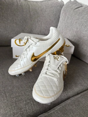 Nike Tiempo Ronaldinho guld/vit - Exklusiva fotbollsskor från Nike Tiempo Ronaldinho-kollektionen i vitt läder med gulddetaljer. Skorna har quiltad tå, guldfärgad Nike Swoosh och guldfärgade dobbar. Klassisk snörning och specialdetaljer med Ronaldinho-signatur. Helt nya och oanvända.