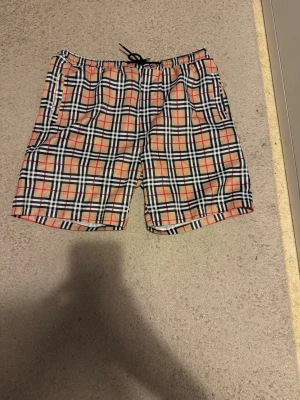 Rutiga shorts med snörning - Säljer ett par rutiga shorts med snörning i midjan. Shortsen har en klassisk beige, svart, vit och röd färgkombination och elastisk midja för skön passform. Perfekta för sommaren och har fickor på sidorna.