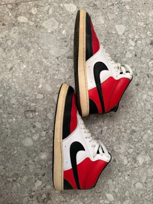 Nike Air Jordan 1 High röd/vit/svart - Klassiska Nike Air Jordan 1 High sneakers i rött, vitt och svart. Skorna har högt skaft, svart swoosh, vita skosnören och en slitstark yttersula. Ovandelen är i läder och designen är ikonisk med färgblock och perforerad tå.