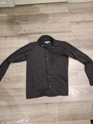 Svart jacka från SMOG med tryckknappar - Stilren svart jacka från SMOG i relaxed fit. Jackan har tryckknappar framtill, klassisk krage och två sidofickor. Materialet är mjukt och lätt, perfekt för lager på lager. Enkel och clean design som passar till det mesta.