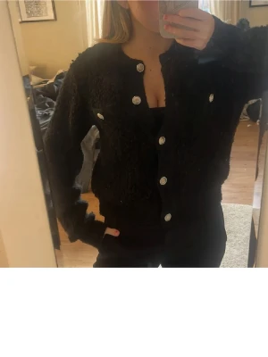 Svart fluffig kofta med silverknappar - Säljer en svart, fluffig kofta med rund hals och stora silvriga knappar framtill. Koftan har två dekorativa bröstfickor och ribbade muddar vid ärmslut och nederkant. Perfekt för dig som gillar en mysig och trendig look.
