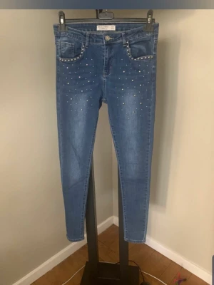 Blå jeans med nitar från Hello Miss - Snygga blå jeans från Hello Miss med silverfärgade nitar längs fickorna och framtill på benen. Jeansen har en klassisk femficksmodell, normal midja och smal passform. Tillverkade i stretchigt denim för extra komfort.