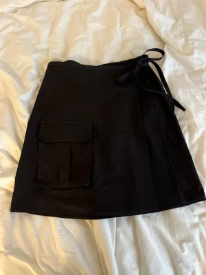 GANNI kjol med ficka och knytband - Stilren svart kjol med  ficka framtill och knytband i midjan. Omlottmodell som ger en cool look och extra komfort. Perfekt för dig som gillar en enkel men trendig stil. Materialet känns mjukt och följsamt. Storlek 40 men passar även 42 skulle jag säga! Tyvärr inte kommit till användning så den är knappt använd. Nypris 2200kr. 