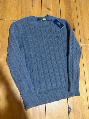 Grå kabelstickad tröja Polo Ralph Lauren - Säljer en grå kabelstickad tröja från Polo Ralph Lauren med rund hals och diskret logga på bröstet. Tröjan har långärmad passform och ribbade muddar vid ärmslut och nederkant. Perfekt för dig som gillar klassisk stil med en modern touch. 