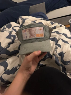 Grå keps med 1000-kronors patch - Cool grå keps med mesh på sidorna och en unik patch framtill som föreställer en svensk 1000-kronorssedel. Perfekt för dig som vill sticka ut och ha något annorlunda på huvudet. Justerbar baktill för bästa passform.