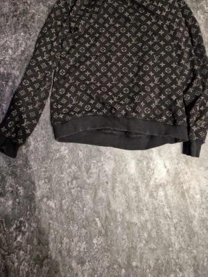 Svart Louis Vuitton sweatshirt - Säljer en svart sweatshirt från Louis Vuitton med det klassiska monogrammönstret i grått över hela tröjan. Tröjan har rund hals, långa ärmar och ribbade muddar vid ärmslut och nederkant. Perfekt för dig som gillar streetwear och exklusiva märken. Vill bli av med snabbt, därför lågt pris.