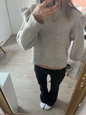 Ljusgrå stickad tröja med volang - Trendig ljusgrå stickad tröja med volangdetaljer över axlarna och lång ärm. Tröjan har en rak, något kortare passform och ribbstickade muddar vid ärmslut och nederkant. Perfekt att styla med jeans eller kjol för en cool vardagslook.