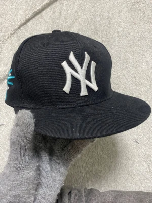 New Era 59FIFTY svart keps New York - Säljer en svart New Era 59FIFTY keps med broderad text 'Greetings from New York' och frihetsgudinnan i turkos och vitt på sidan. Platt skärm i ljus beige och klassisk passform. MLB Genuine Merchandise. Perfekt för dig som gillar streetwear och snygga detaljer.