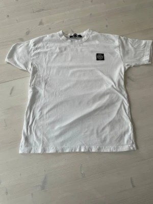 Vit Stone Island t-shirt i bomull - Säljer en clean vit t-shirt från Stone Island med klassisk logopatch på bröstet. T-shirten har rund hals och korta ärmar, perfekt till alla outfits. Tillverkad i mjuk bomull för skön känsla hela dagen. Storlek 176. Är lite liten i storlek så den passar folk med ett snäpp under 
