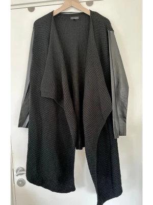 Svart kofta med ärmar i skinnimitation  - Supersnygg svart stickad kofta med långa ärmar i läderimitation. Från Kappahl i storlek 44/46/L/XL. Koftan har en öppen design utan knäppning och en assymetrisk nederkant med vackert fall. Kan bäras som den är eller med skärp i midjan. Fint skick!  Skickas mot porto, kan även hämtas i Stockholm.