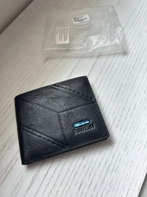 Svart läderplånbok från Boweisi - Snygg svart plånbok från Boweisi i äkta läder. Plånboken har modern design med geometriska sömmar, flera kortfack, myntfack med dragkedja och plats för ID-kort. Perfekt för dig som vill ha stilren och praktisk accessoar.