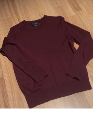 Mörkröd stickad tröja från Riley - Säljer en enkel och stilren mörkröd stickad tröja från Riley. Tröjan har rund halsringning och långa ärmar, perfekt för lager-på-lager. Materialet är mjukt och bekvämt, och färgen är djup burgundy. Passar dig som gillar klassiska plagg med en modern touch.