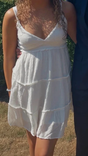 Vit volang klänning, studentklänning  - Söt vit kortklänning med omlott och v-ringad front. Klänningen har tunna axelband och flera volanglager som ger ett luftigt och somrigt intryck. Perfekt för varma dagar och enkel att matcha med accessoarer. Materialet känns lätt och bekvämt. Perfekt till studenten, skolavslutningen, konfirmationen och sommaren!