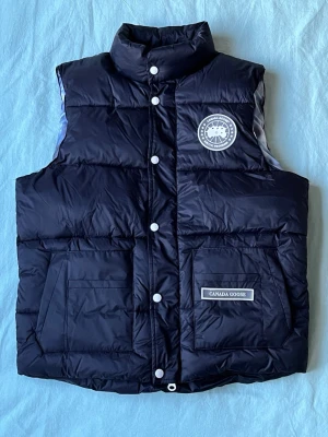 Svart dunväst från Canada Goose - Snygg svart dunväst från Canada Goose med hög krage, stora framfickor och tydlig logotyp på bröstet. Västen har tryckknappar framtill och klassisk quiltad design. Perfekt för lager-på-lager och riktigt varm tack vare dunfyllningen.