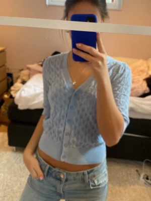 Ljusblå kort kofta med mönster - Säljer en ljusblå, kort kofta med v-ringning och knappar framtill. Koftan har ett diskret stickat mönster och är tillverkad i ett tunt, mjukt material. Perfekt att slänga över en topp för en chill look.