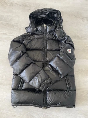 Svart dunjacka från Moncler - Snygg svart dunjacka från Moncler med glansig finish och klassisk pufferdesign. Jackan har huva, två dragkedjeförsedda fickor och Moncler-logga på ärmen. Perfekt för kalla dagar och ger en riktigt trendig look.