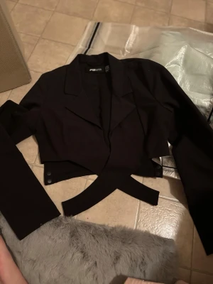 Svart croppad bolerotopp FB Sister XS - Snygg svart croppad bolerotopp från FB Sister i storlek XS. Toppen har djup v-ringning, omlott-design och långa ärmar. Stilren krage och figurnära passform som ger en cool och trendig look. Perfekt för dig som gillar att sticka ut med din outfit.